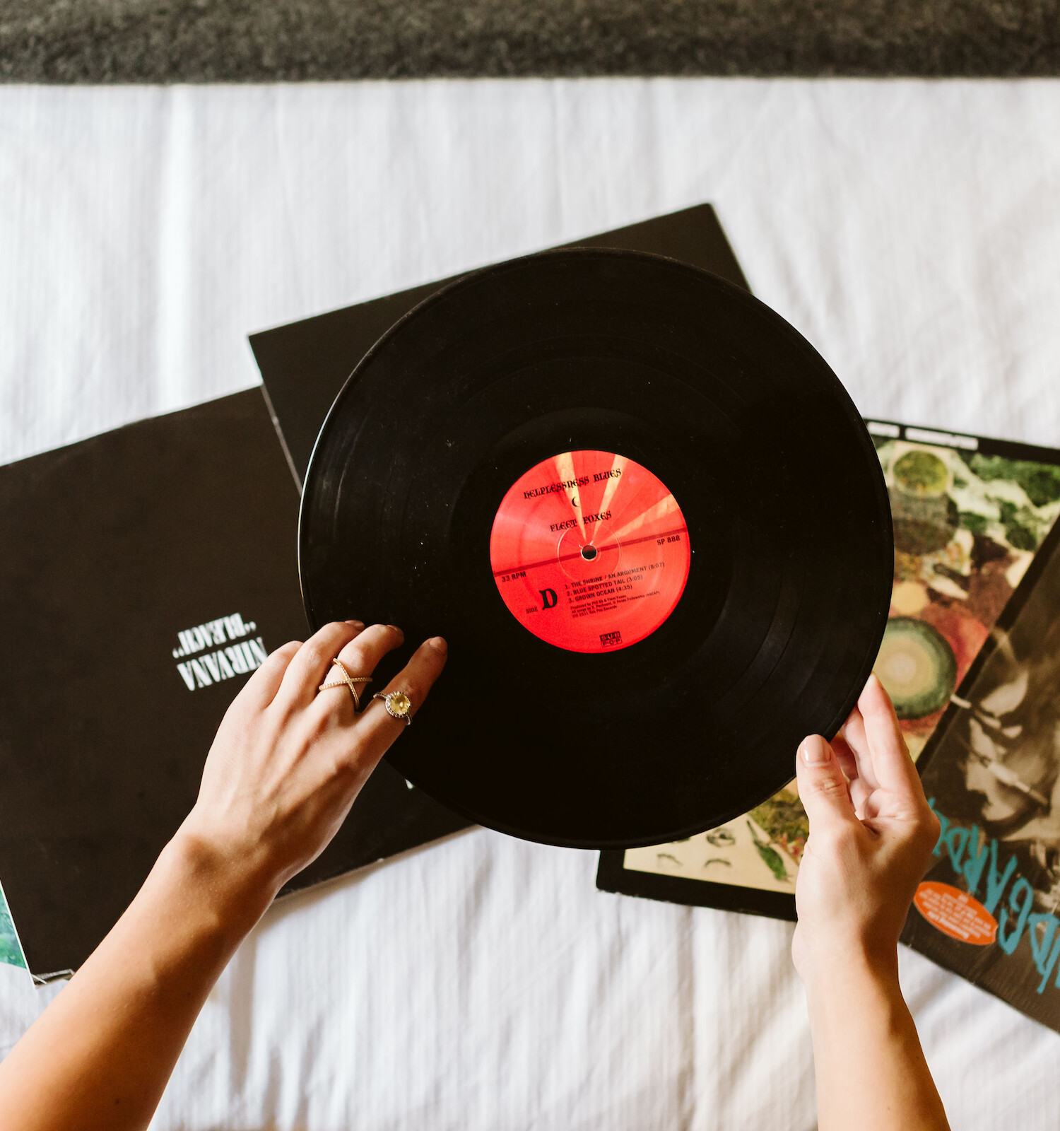 Someone is holding a vinyl record above several other record covers on a white surface.