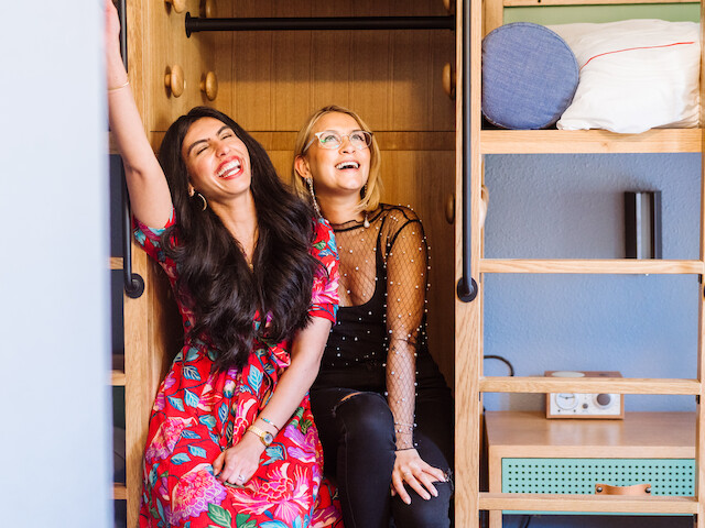 Two people are sitting and laughing in a cheerful, cozy space with a wooden backdrop and shelves with various items.