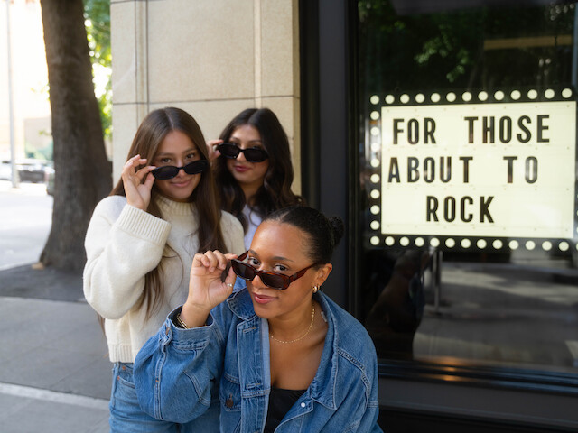 Three people pose with sunglasses near a sign that reads 