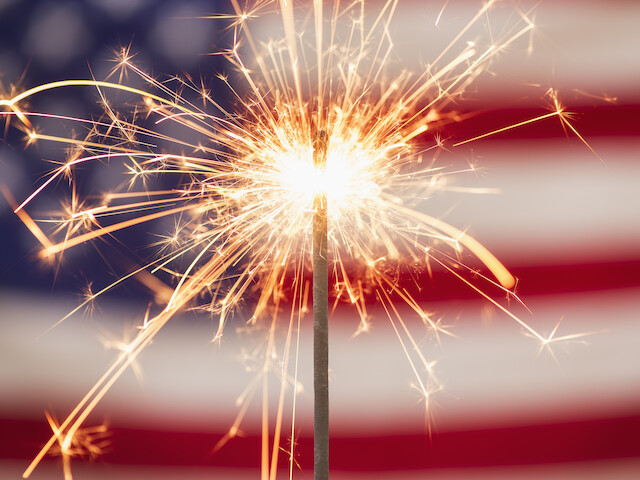 A sparkler is lit in the foreground with a blurred American flag in the background.