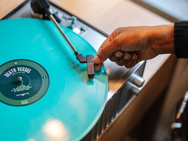 A person is placing a stylus on a spinning turquoise vinyl record, set on a turntable by a window, creating a cozy atmosphere.