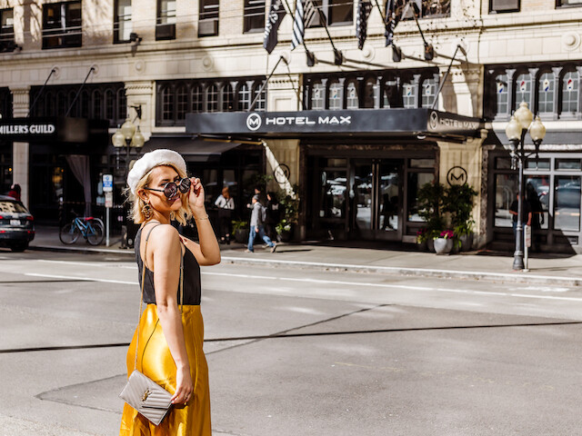A stylish person in sunglasses and a yellow skirt stands on a city street near Hotel Max, with a purse in hand.