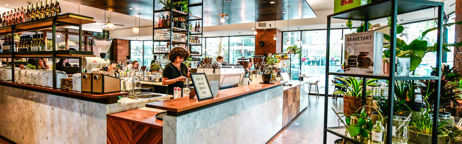 The image shows a modern cafe interior with a counter, shelves of plants, and a warm, inviting atmosphere.