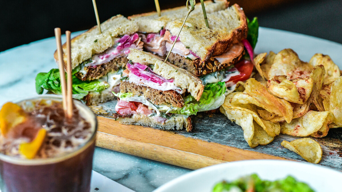 A sandwich with lettuce and tomatoes, served with chips and a beverage, is placed on a wooden board.