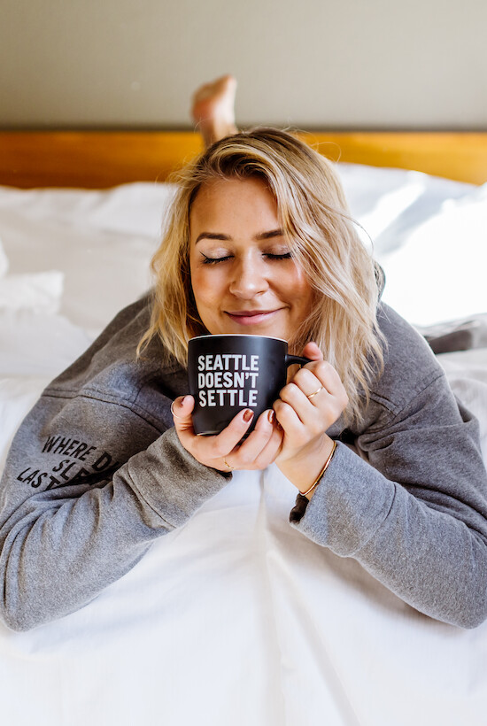 A person lies on a bed, holding a mug that says 