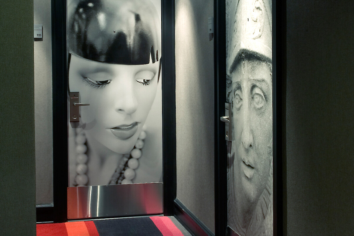 A hallway with two doors featuring large black-and-white photos, one of a woman and one of a statue, on a striped carpet floor.