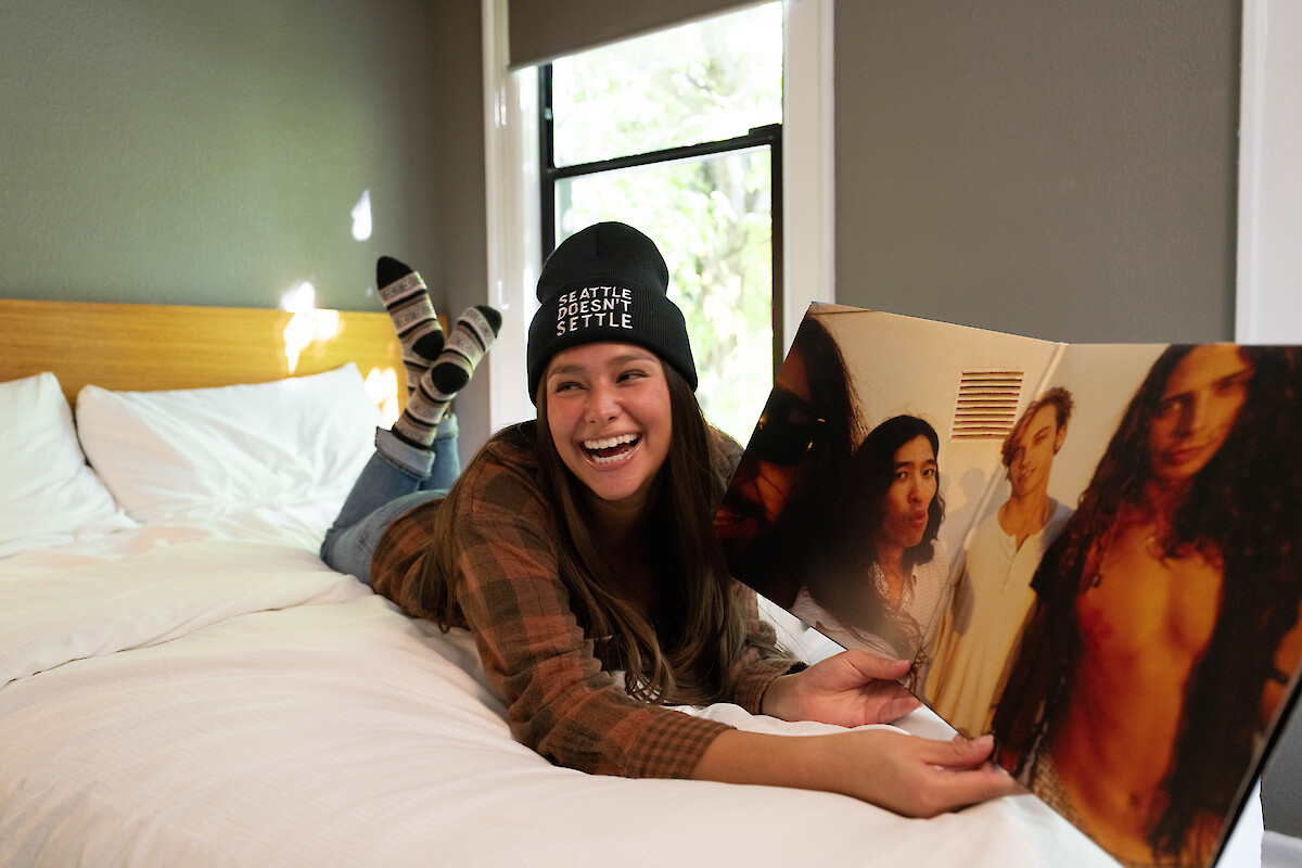 A person lies on a bed, smiling and holding open a vinyl record jacket while wearing a beanie that reads 