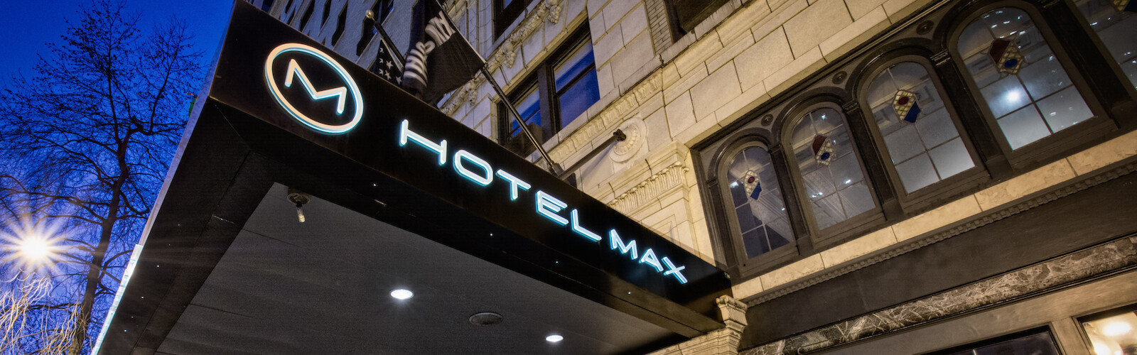 The image shows the entrance of Hotel Max with a prominent sign, located in a city setting with surrounding buildings.