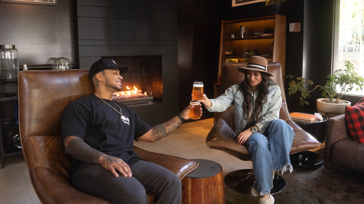 Two people in a cozy room with a fireplace, sharing a toast while sitting in leather chairs.
