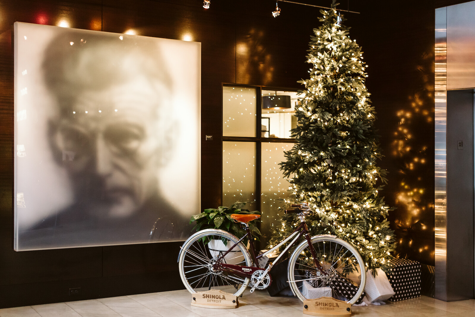 A cozy holiday scene: a tall Christmas tree with warm lights beside two vintage bicycles, in front of a large blurred portrait mural.