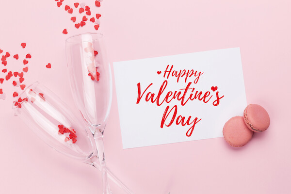 Two champagne flutes with red heart confetti, a “Happy Valentine’s Day” card, and pink macarons on a pink background.