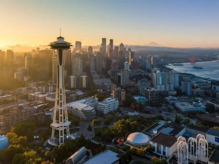 Aerial view of Seattle at sunset featuring the Space Needle, a mix of skyscrapers, water to the right, and warm golden light washing over the city.