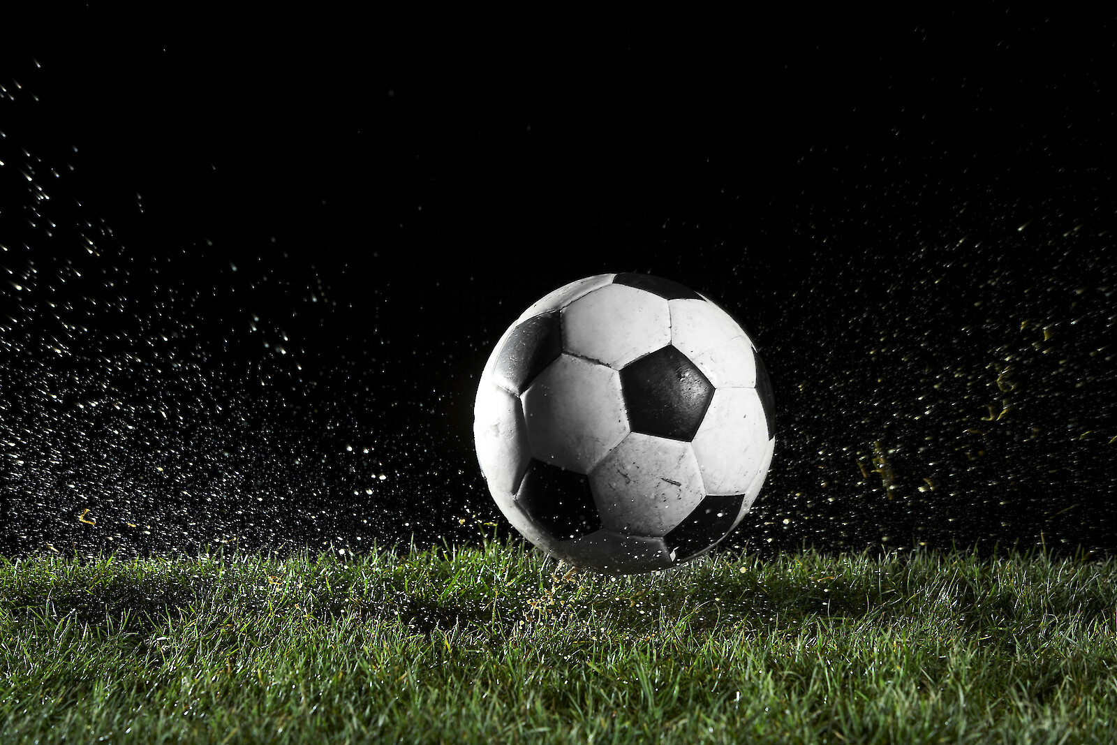 A black-and-white soccer ball sits on a grassy field with soil spray around it as droplets freeze midair, dramatic night lighting, dynamic sports vibe.