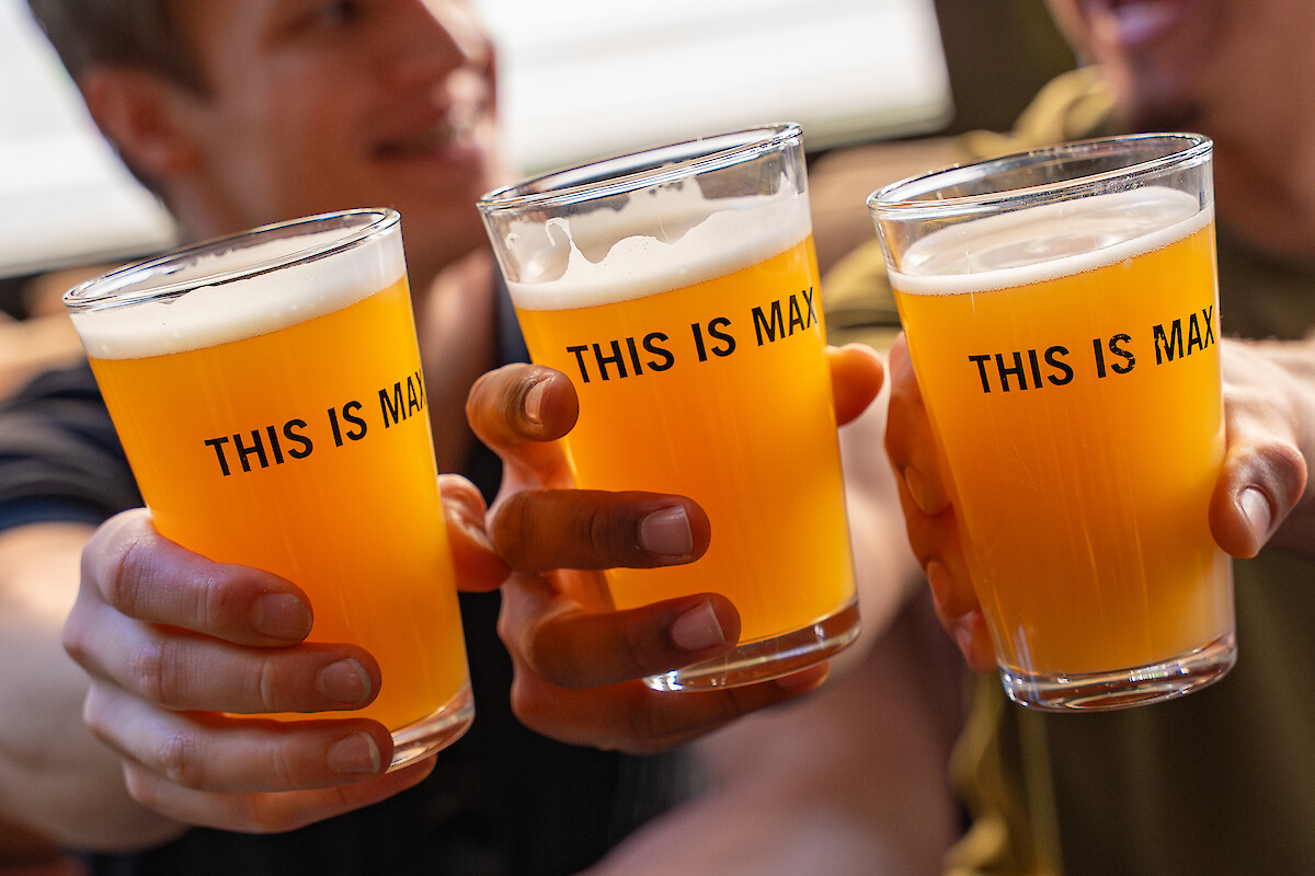 Three friends toast with orange drinks labeled “THIS IS MA,” smiling at a cozy hangout.