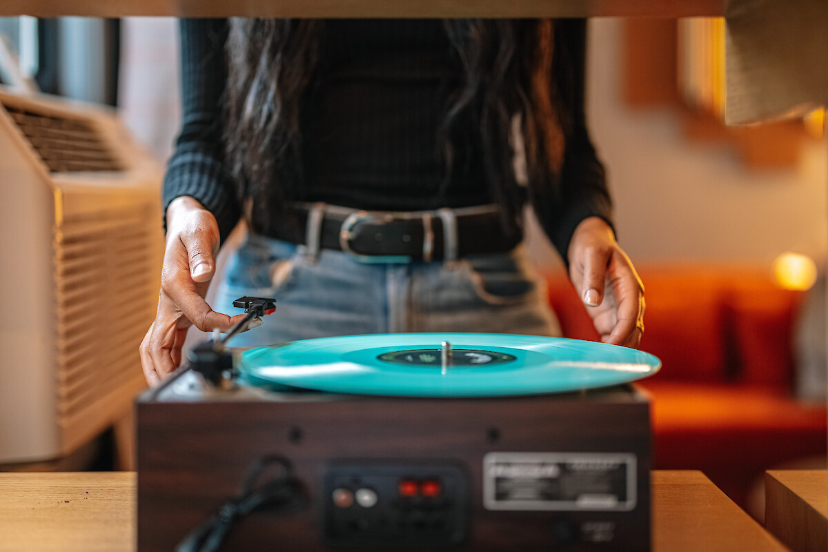A person is spinning a turquoise vinyl on a retro turntable; hands on the tonearm, cozy room in the background, warm lighting.