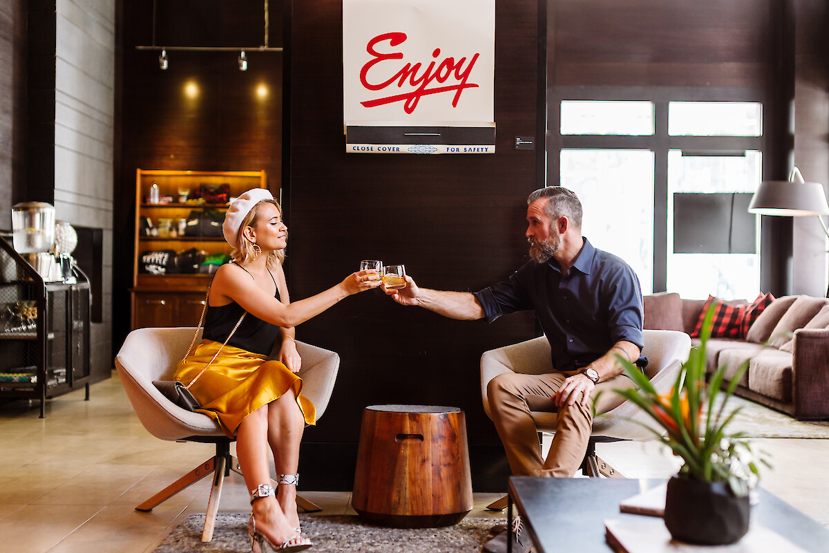Two people toast a drink in a stylish living room; a modern, cozy lounge with plants, warm wood tones, and a sign that reads “Enjoy.”