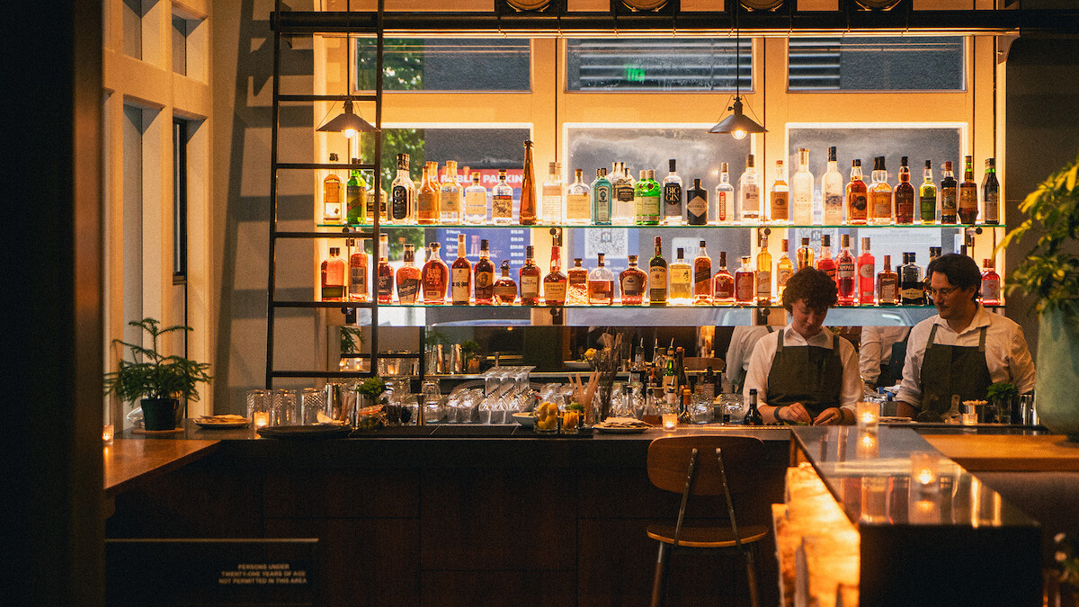 A cozy bar with two bartenders preparing drinks, illuminated by warm lighting, and a variety of liquor bottles displayed on shelves.