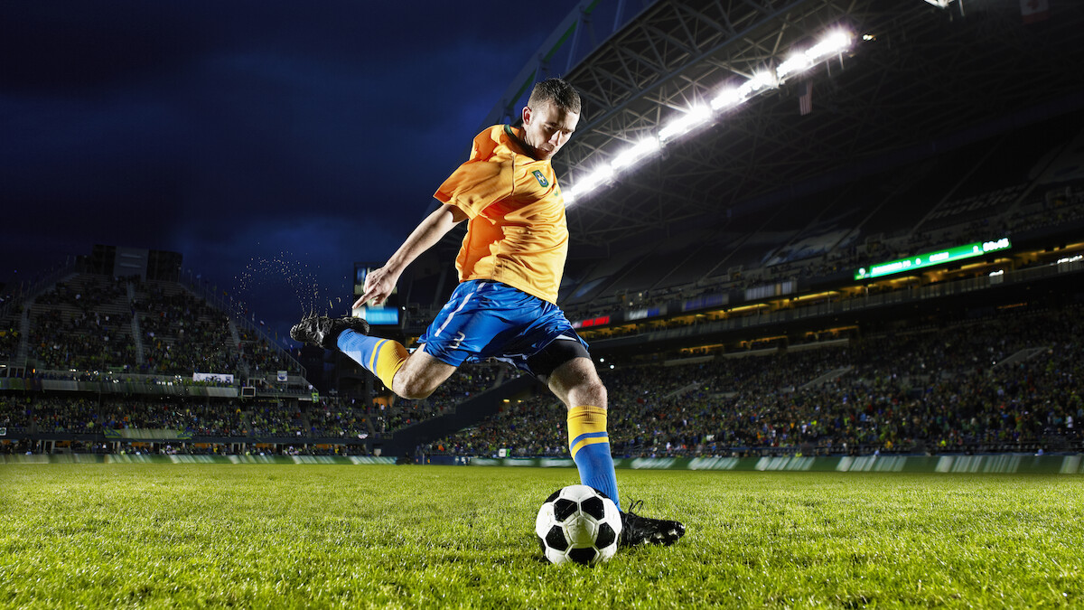 A soccer player in an orange jersey and blue shorts kicks a black-and-white ball on a lit, grassy stadium field at night, mid-action.