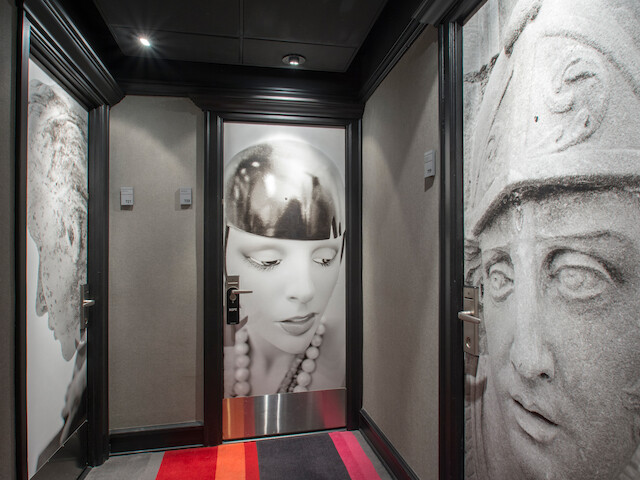 The image shows a hallway with black framed doors decorated with large black-and-white portraits of a woman and stone sculptures of faces on the walls.