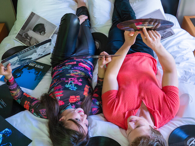 Two friends lie on a bed, surrounded by vinyl records and album covers, enjoying a relaxed music and conversation session.