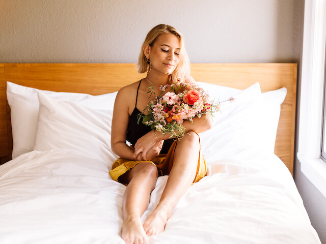 A woman in a black top and yellow shorts sits on a bed holding a colorful bouquet in a well-lit room.