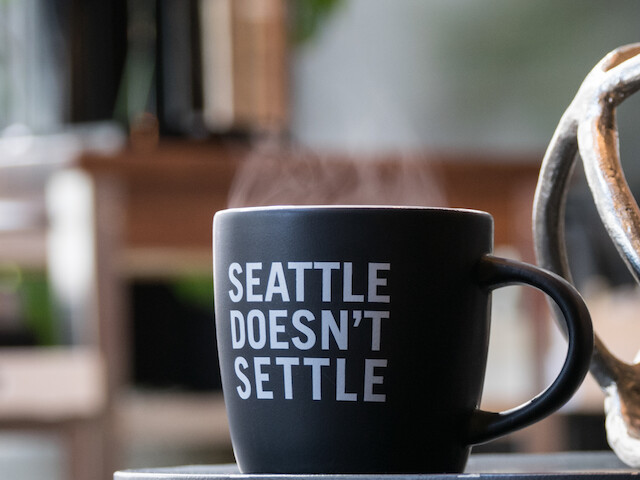A black mug with white text “SEATTLE DOESN'T SETTLE” on a table, blurred kitchen scene in the background, all in focus.