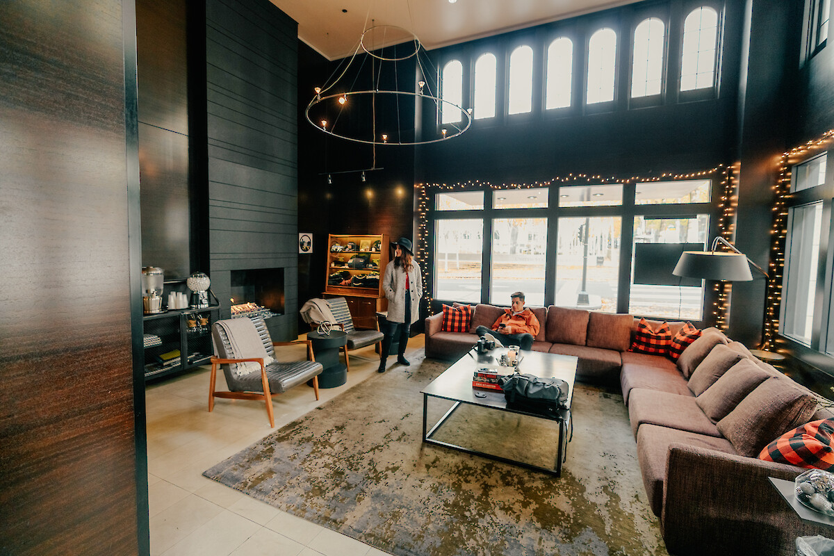 A cozy, modern living room with large windows, festive string lights, a sectional sofa, and two people relaxing and chatting.