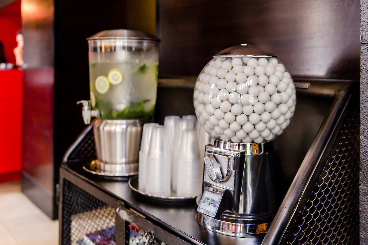 The image shows a beverage station with a juice dispenser, a bowl of white gumballs, and stacked cups on a black counter.