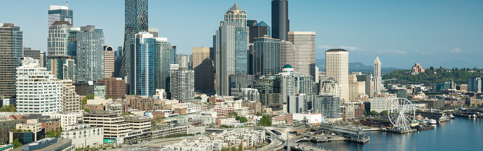 A city skyline with high-rise buildings, a waterfront, a pier, and a Ferris wheel under a clear blue sky.