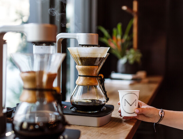 A person brews coffee with pour-over equipment, holding a paper cup with a logo on it.