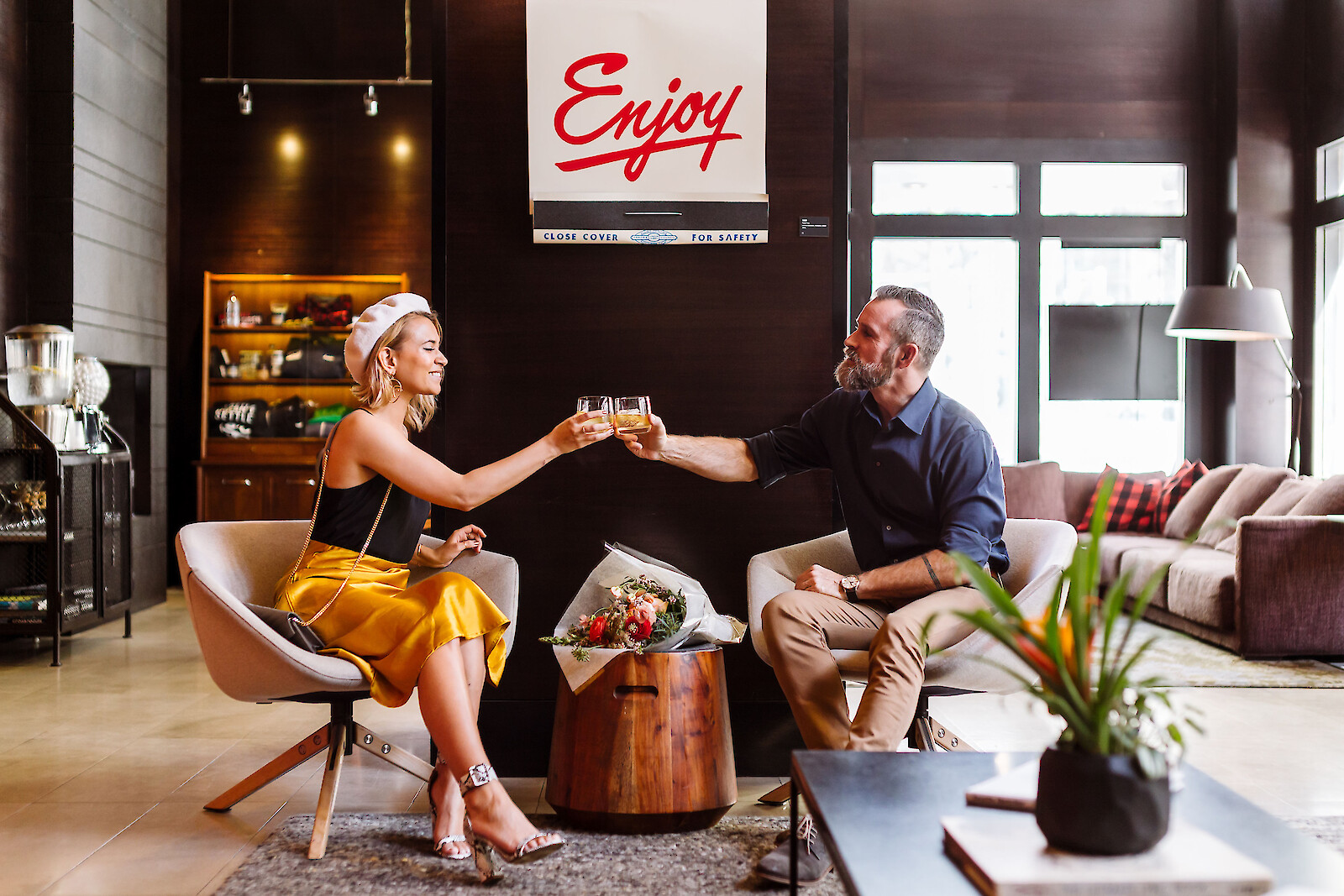 A couple toasting drinks in a cozy, stylish living room with modern décor and a bright “Enjoy” sign, capturing the warm, welcoming vibe of Hotel Max’s Black Friday 2025 and Cyber Monday cyber sale.