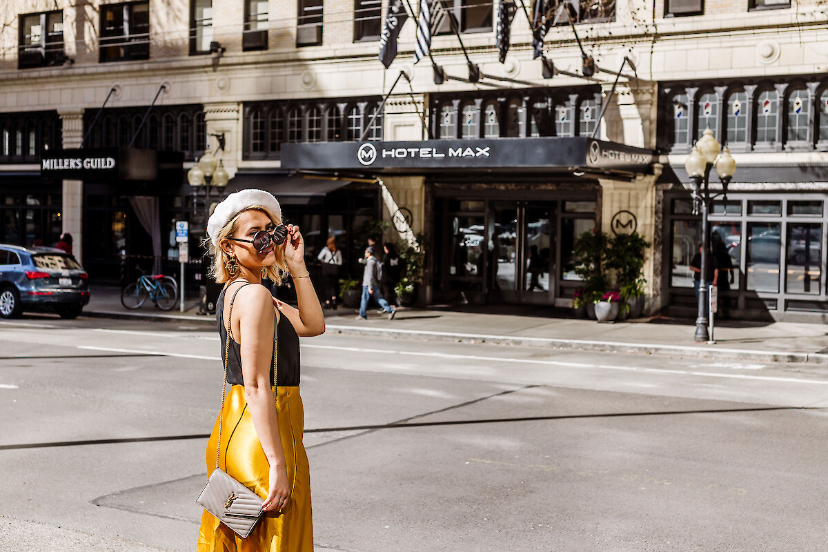 A stylish woman in yellow pants and black top stands on a city street, wearing round sunglasses and a beret, with the Hotel Max in the background.