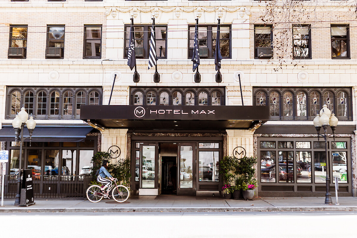 A woman rides a bicycle past the Hotel Max, a building with black-and-white flags, plants, and large windows on an urban street.