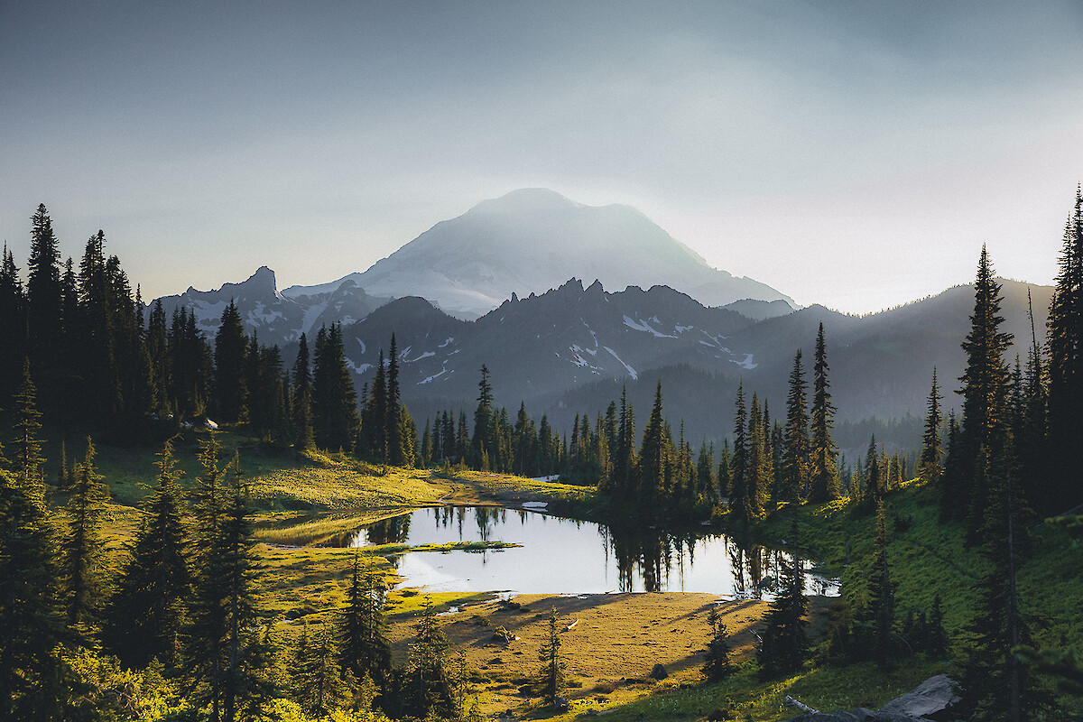 A scenic mountain landscape with a lake, dense trees, and snow-capped peaks in the background, bathed in soft sunlight.