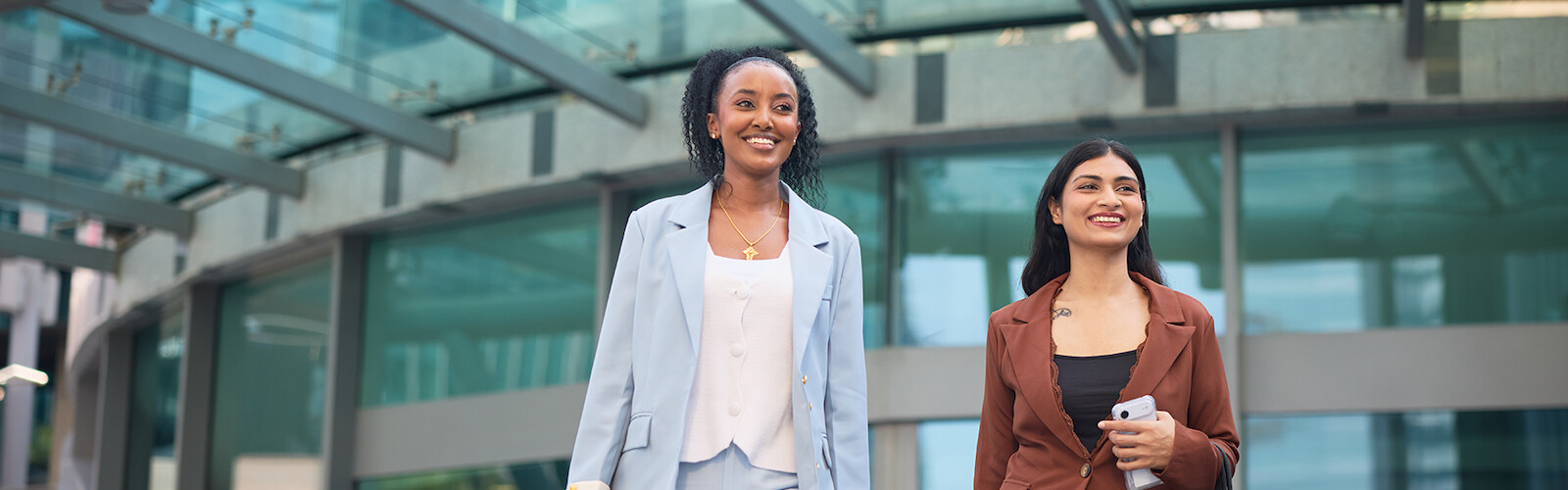 Two professional women walking outside a modern building, smiling and carrying handbags and a suitcase.