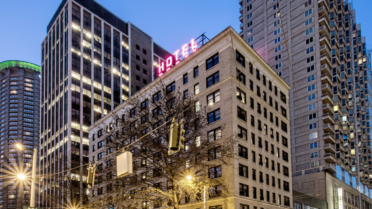 A city street scene at dusk with tall buildings, a hotel sign on top, streetlights, and cars moving through the intersection.