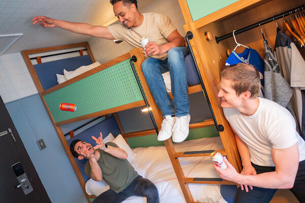Three friends are having fun on bunk beds in a hotel room, playing and laughing with snacks and drinks.