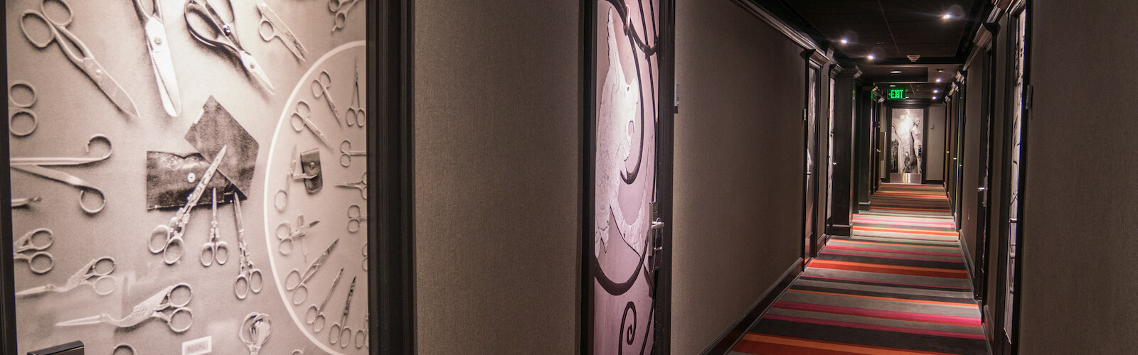 A hotel hallway with a door featuring scissors and sewing tools, colorful carpeting, and an exit sign at the end.