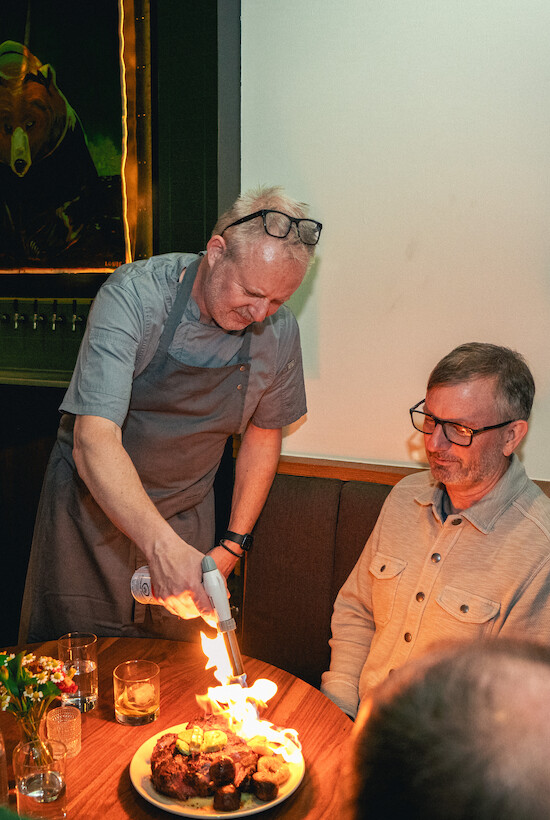 A chef performs a flaming presentation at a dinner, entertaining a guest sitting at the table with a plate of food.