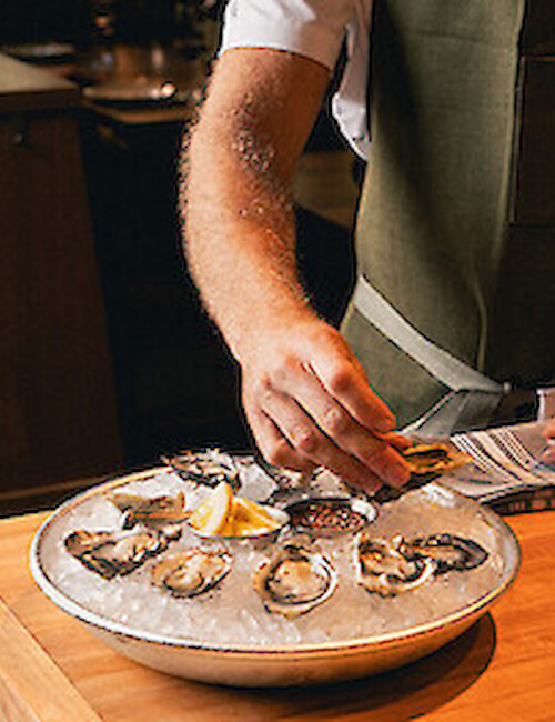 Person prepares fresh oysters on a wooden tray, adding lemon slices in a cozy, upscale seafood setting.