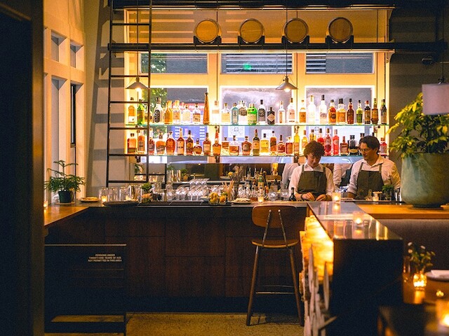 A bar scene with two bartenders behind the counter, bottles of liquor on shelves, warm lighting, and cozy ambiance.