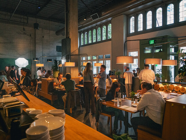 People dining and socializing inside a cozy, well-lit restaurant with large windows and hanging lamps.