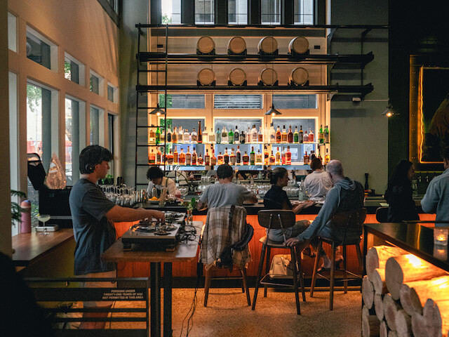 A lively bar with a high ceiling, large windows, wooden barrels, and patrons enjoying drinks in a cozy, modern ambiance.