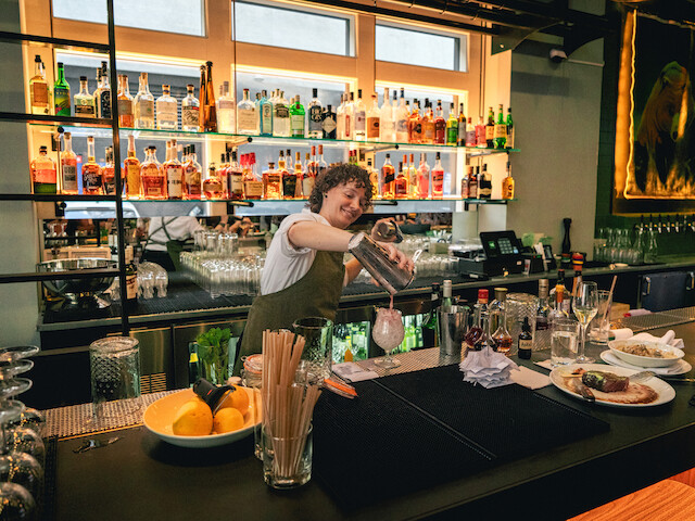 A bartender with curly hair pours a drink at a lively bar, surrounded by bottles, glasses, and snacks in a modern, vibrant setting.