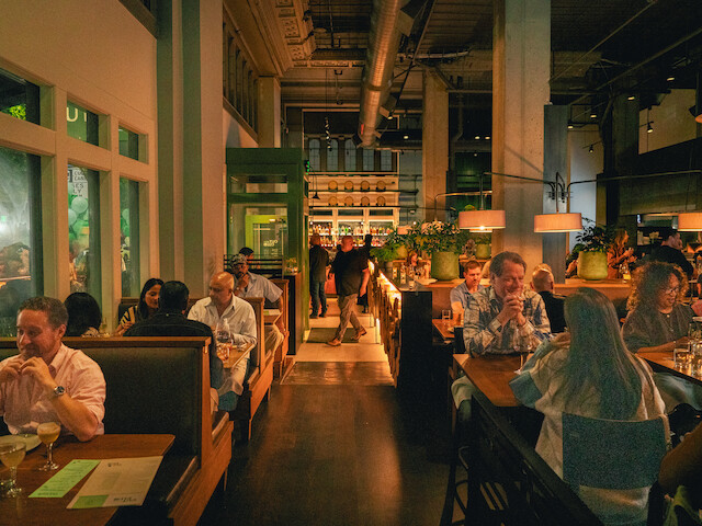 A lively restaurant interior with people dining, warm lighting, greenery, and a bar area in the background.