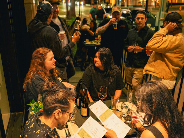 People are dining and socializing outside a restaurant at night, with some holding drinks and reviewing menus.