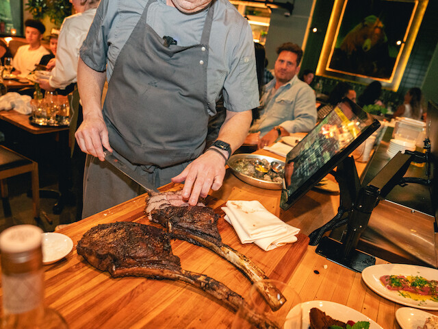 A chef is slicing large grilled steaks at a lively restaurant with diners watching and colorful decor in the background.