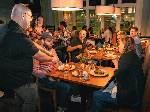A waiter sprays something on a seated man's food while others around them enjoy dinner at a lively restaurant.