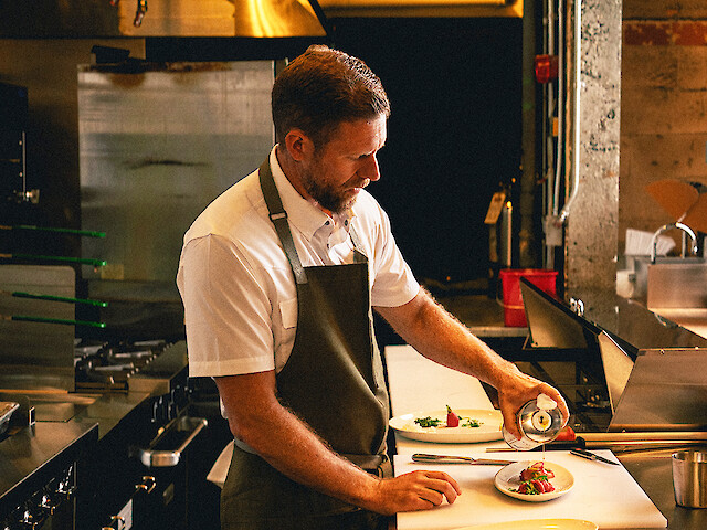 A chef is plating a dish in a professional kitchen, pouring sauce onto a beautifully prepared plate of food.