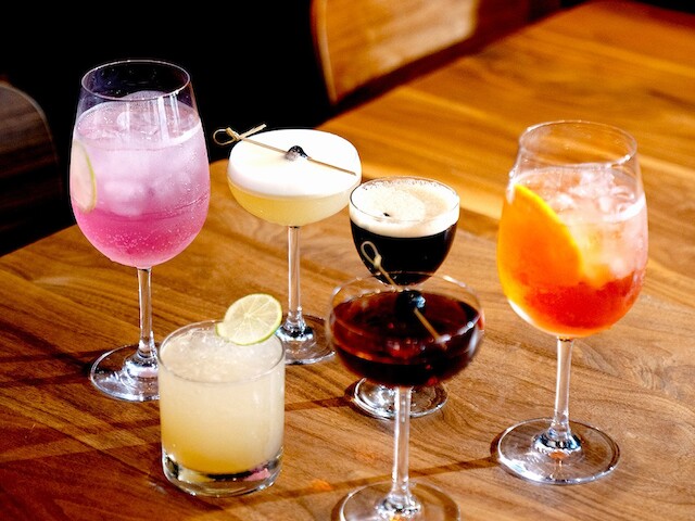 A variety of colorful cocktails and drinks are displayed on a wooden table, showcasing different styles and garnishes.